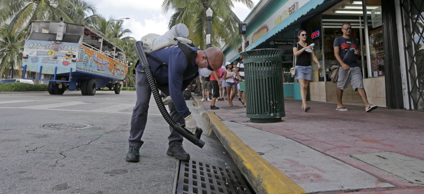 Miami-Dade mosquito control inspector Yasser "Jazz" Compagines sprays a chemical mist into a storm drain, as a tour vessel passes by at left, Tuesday, Aug. 23, 2016, in Miami Beach, Fla.