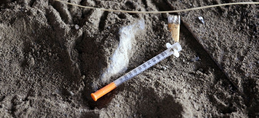 a discarded syringe sits in the dirt with other debris under a highway overpass. 