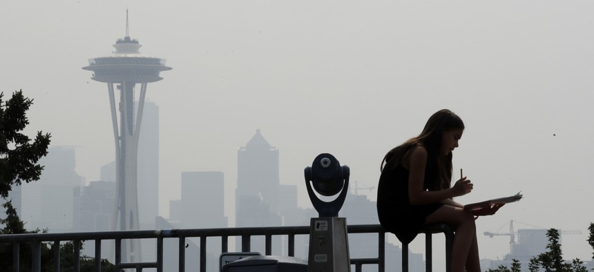 A girl works on a drawing next to an unused viewing scope as a smoky haze from wildfires obscures the Space Needle and downtown Seattle behind, Tuesday, Aug. 14, 2018.