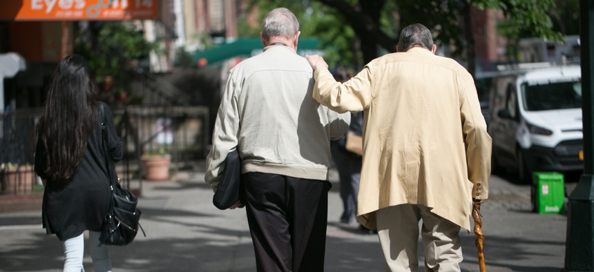  Two elderly men help each other walk down a street.