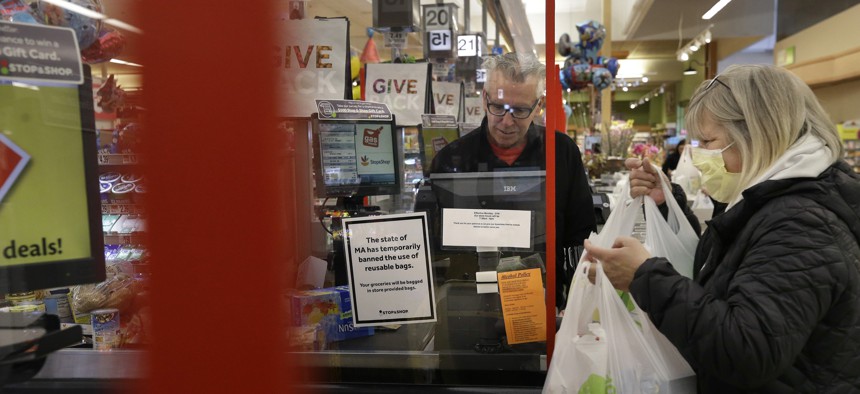 A cashier at a grocery store in Quincy, Mass., center, works behind a plastic shield as a shopper, right, places groceries in a cart.