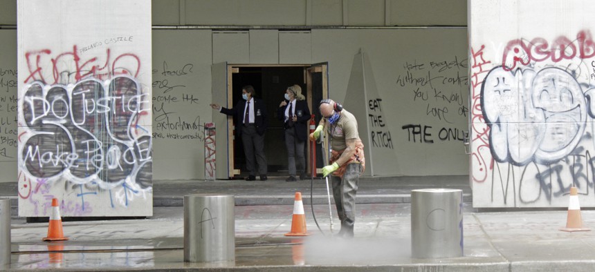 In this July 8, 2020 photo, a worker washes graffiti off the sidewalk in front of the Mark O. Hatfield Federal Courthouse in downtown Portland, Ore., as two agents with the U.S. Marshals Service emerge from the boarded-up main entrance.