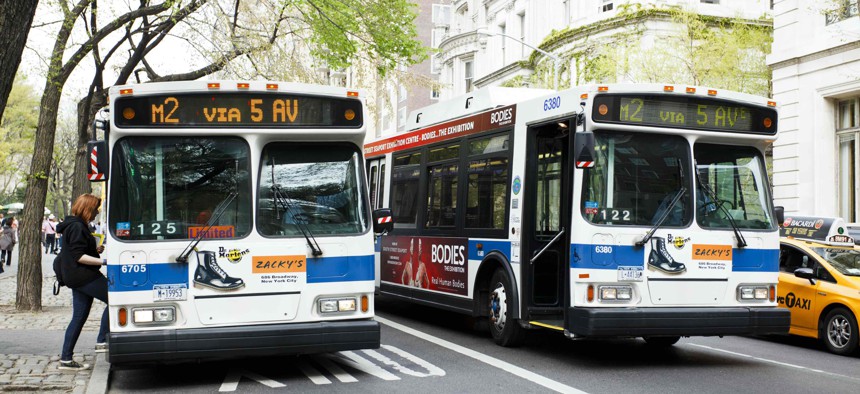 Two buses side by side on Fifth Avenue in Manhattan. One bus is stopped and taking on a passenger. 