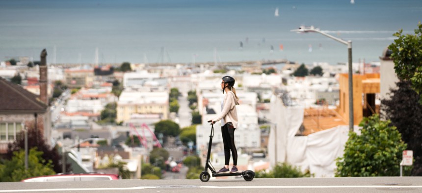 Woman safely riding an e-scooter with a helmet with the San Francisco Bay in the background.