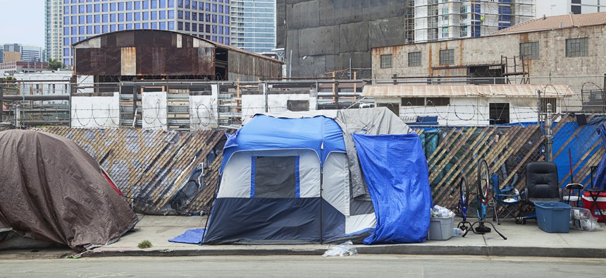 Tents and gear of a homeless encampment along a street with industrial buildings and skyscrapers in the background on Aug. 20, 2021. 