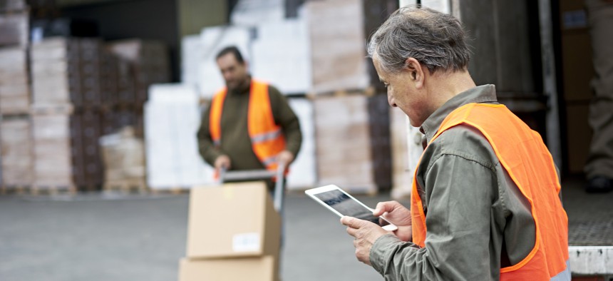 A man looks at a tablet as another worker moves boxes from a truck.