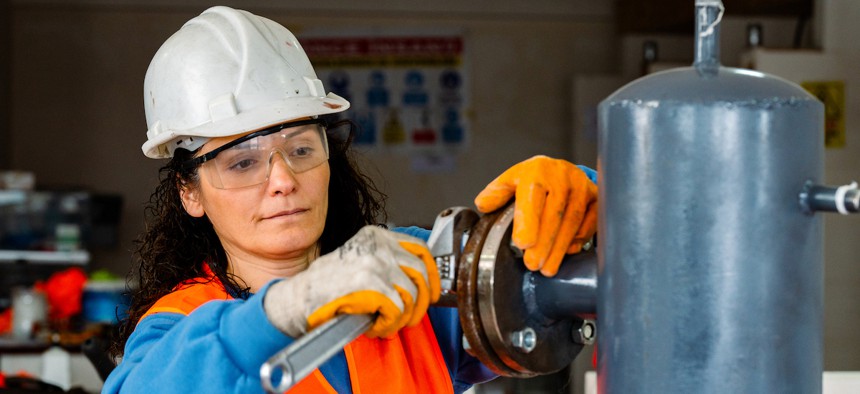 Woman wearing a white hard hat screws a wrench on machinery.