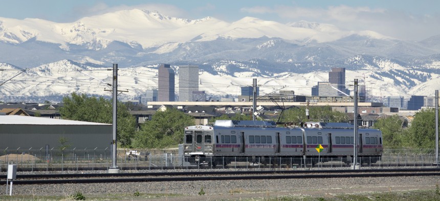 A commuter rail rolls down the tracks past downtown Denver skyscrapers framed by snowy Mount Evans and the Rocky Mountains.
