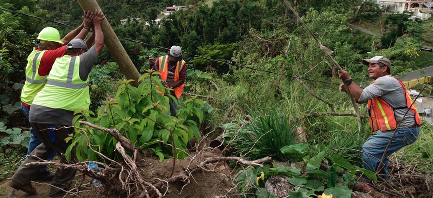 Public Works Sub-Director Ramon Mendez, wearing a hard hat at left, works with local municipal workers to install a new electrical post to return power to a home without any, four months after Hurricane Maria hit Puerto Rico.