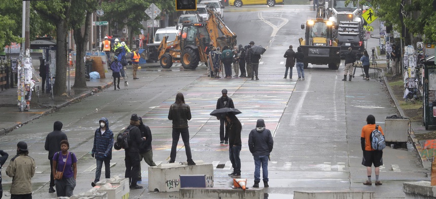 Protesters stand on barricades a block away as Seattle Department of Transportation workers remove other barricades Tuesday, June 30, 2020 at the CHOP (Capitol Hill Occupied Protest) zone in Seattle.
