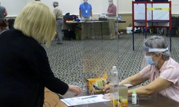 Election worker Adonlie DeRoche, seated, wears a mask and face shield behind Plexiglas for safety while handing a ballot and single-use pen to a voter during the July 14 primary election in Portland, Maine.