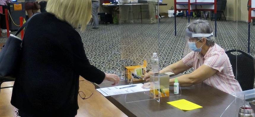 Election worker Adonlie DeRoche, seated, wears a mask and face shield behind Plexiglas for safety while handing a ballot and single-use pen to a voter during the July 14 primary election in Portland, Maine.