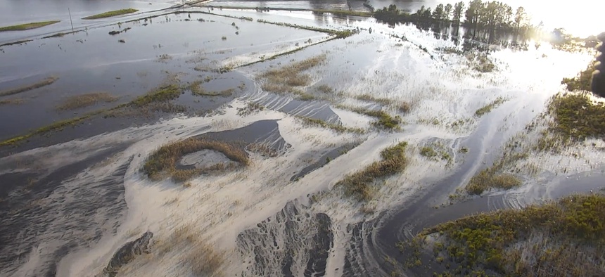 In this image made from 2018 drone video provided by the North Carolina DEQ, light gray material flows out of a flooded coal ash dump toward the Cape Fear River at Duke Energy's L.V. Sutton Power Station near Wilmington, N.C.